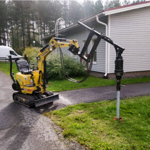 En liten gul Yanmar grävmaskin med en borrutrustning och monterar skruvpålar, arbetar nära ett bostadshus.