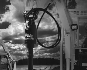 En svartvit bild av en Digga PD Generation borraggregat monterad på en grävmaskin mot en bakgrund av ett öppet fält och molnig himmel.