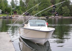 En vit Utility 23 motorbåt förtöjd med Mooring Whips på en brygga vid en lugn sjö, med trähus i bakgrunden.