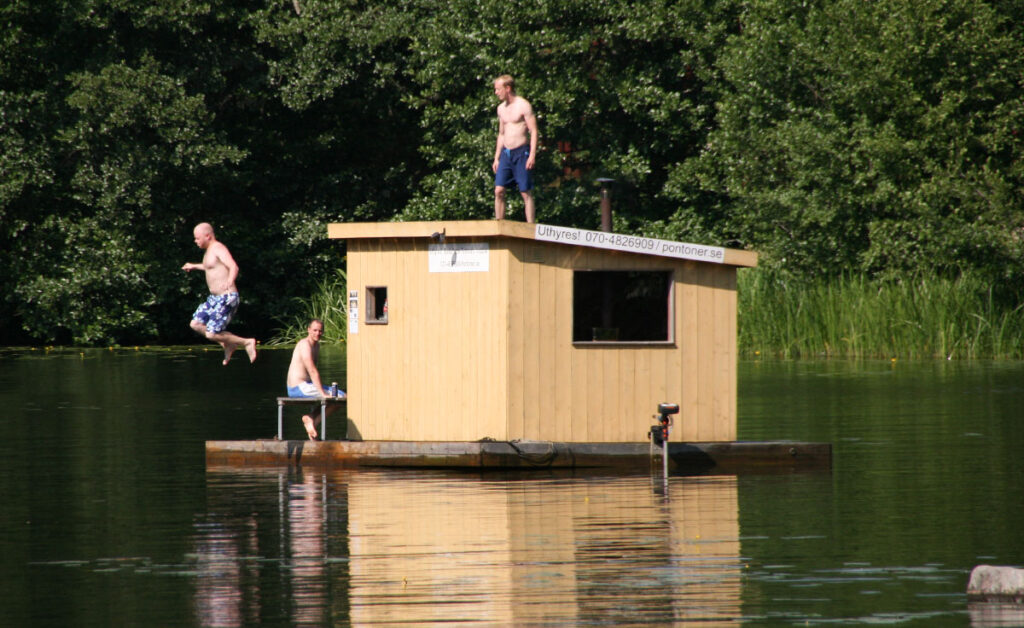 Liten bastuflotte på pontoner med personer som hoppar i vattnet från taket